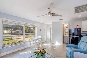 A spacious living room with a ceiling fan, a dining area with chairs, a kitchen with a refrigerator, and a view through large windows. - Dog-friendly home plus attached studio with fenced yard, washer/dryers, & AC (Naples)