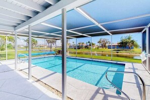 A covered swimming pool with clear blue water, surrounded by a white roof structure, and a view of a residential area with houses and greenery. - Bright Florida home with pool, gas grill, private pool, and washer/dryer (Rotonda West)