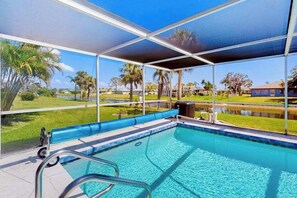 A swimming pool with a covered area, clear blue water, and a view of a grassy area with trees and a body of water. - Bright Florida home with pool, gas grill, private pool, and washer/dryer (Rotonda West)