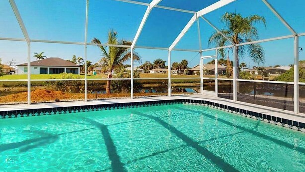 A swimming pool with a clear blue water, surrounded by a white metal structure, with palm trees and houses in the background. - Palm-lined waterfront retreat with enclosed pool, inviting patio, & central AC (Rotonda West)