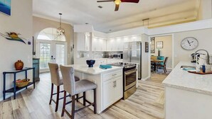 Una cocina moderna con armarios blancos, una isla central, un ventilador de techo y un comedor con mesa y sillas.