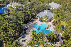 An aerial view of a resort with a central swimming pool, surrounded by palm trees and a small island feature.