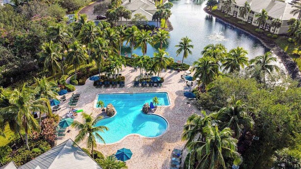 An aerial view of a resort with a central swimming pool, palm trees, and a river.