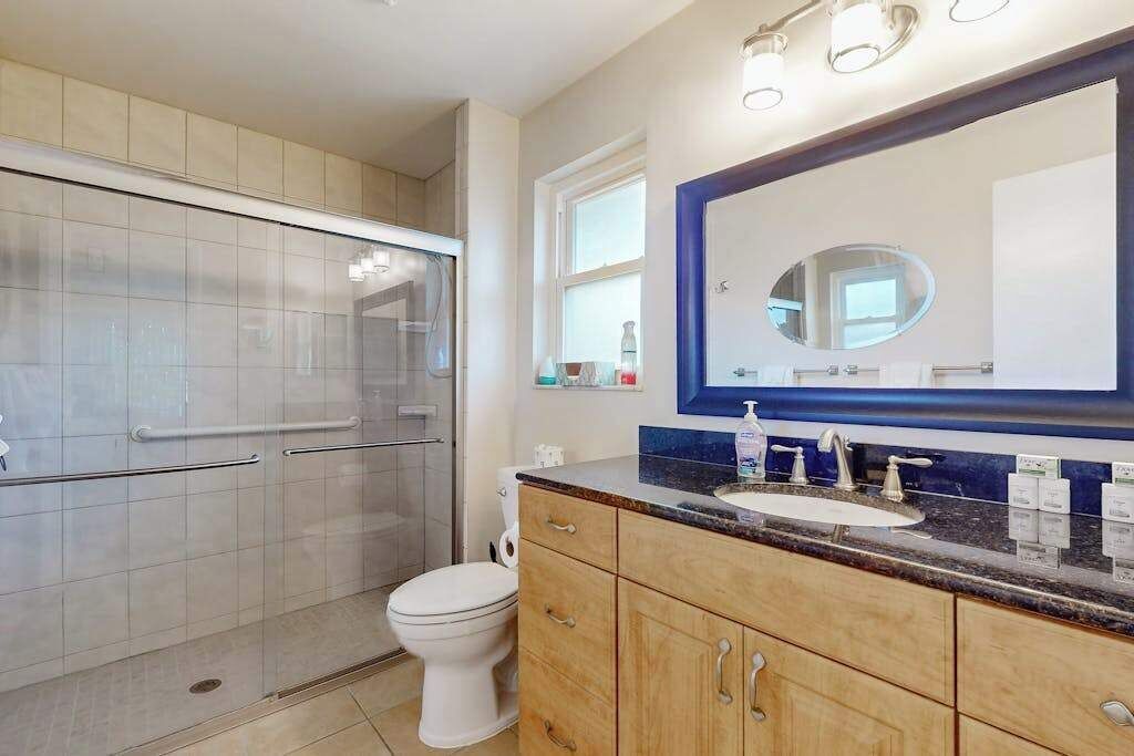 A bathroom with a glass shower enclosure, a white toilet, wooden cabinets, a large mirror, and a granite countertop.