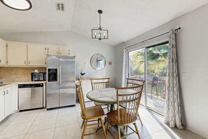 A kitchen with a refrigerator, wooden chairs, a dining table, and a large window with curtains. - Coastal Snowbird Retreat | 30+ Days (Port Charlotte)
