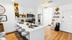 Una cucina moderna con mobili bianchi, un'isola centrale con sgabelli, un forno a microonde, un frigorifero e una decorazione a tema caffè.