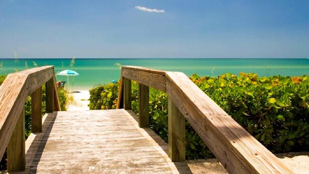 Una passerella in legno che conduce a una spiaggia con acque turchesi cristalline e un ombrellone blu.