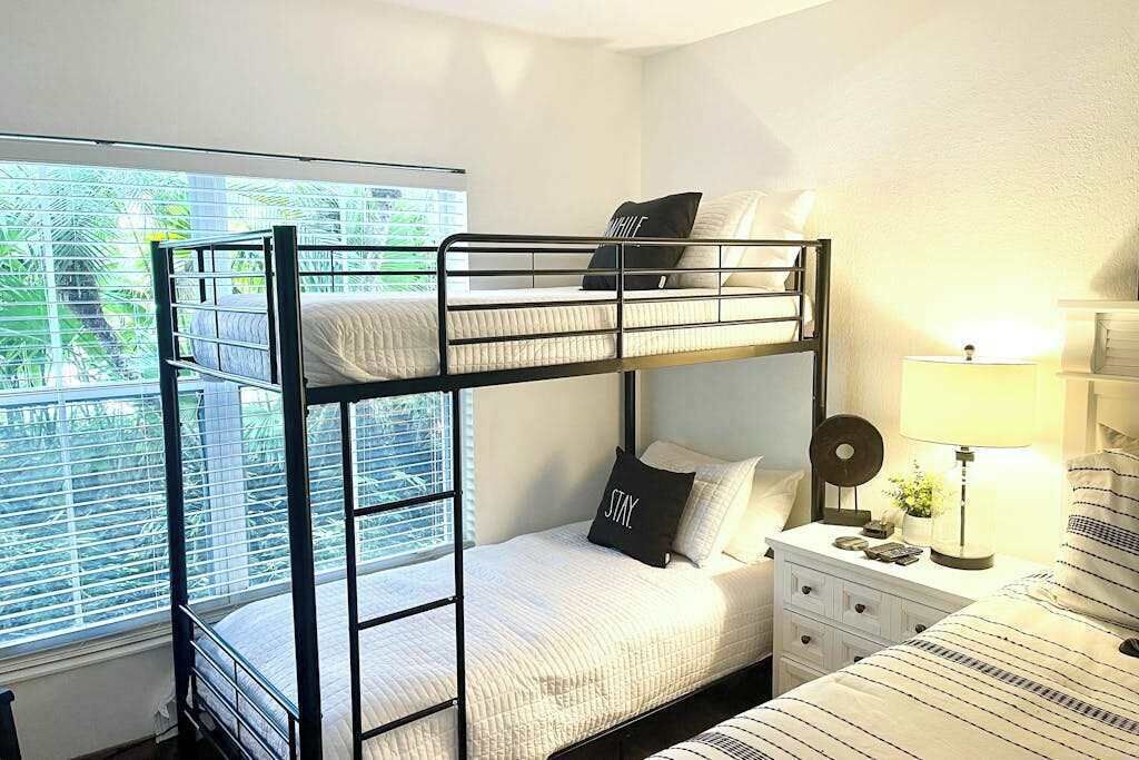 A bunk bed room with a window, a bedside table with a lamp, and a bed with striped bedding.