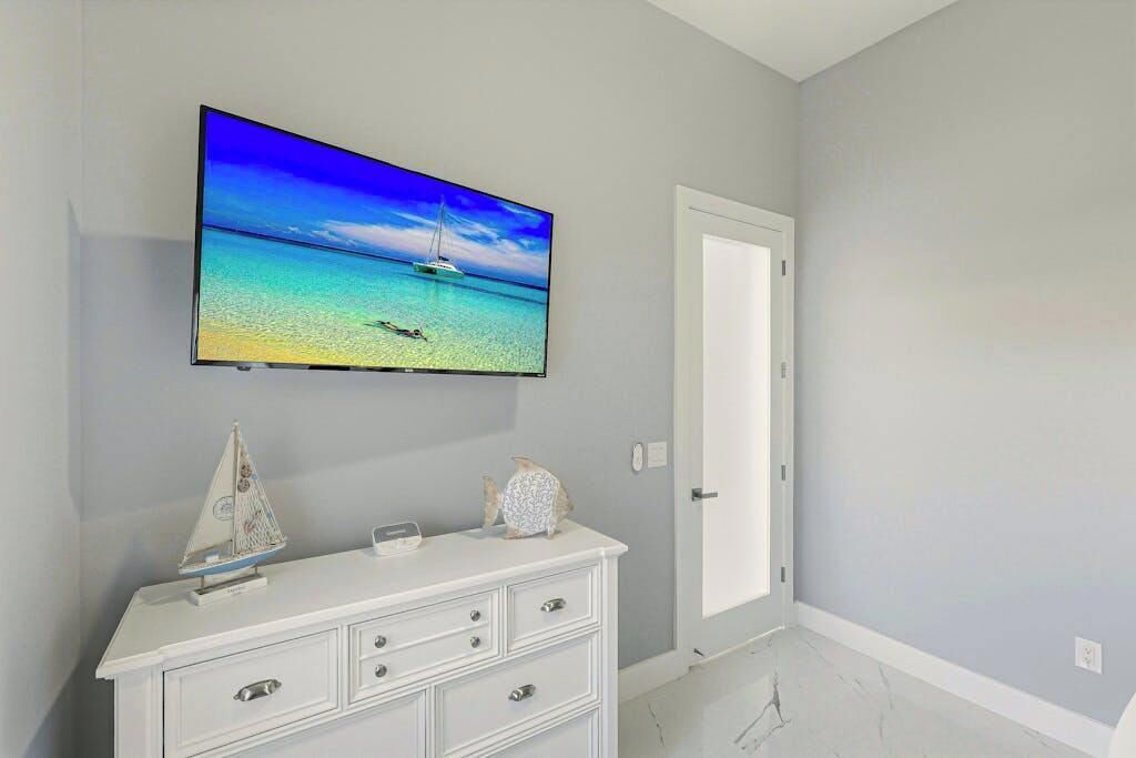 Une pièce avec un téléviseur fixé au mur qui diffuse une scène de plage, une commode blanche avec des tiroirs et une porte.