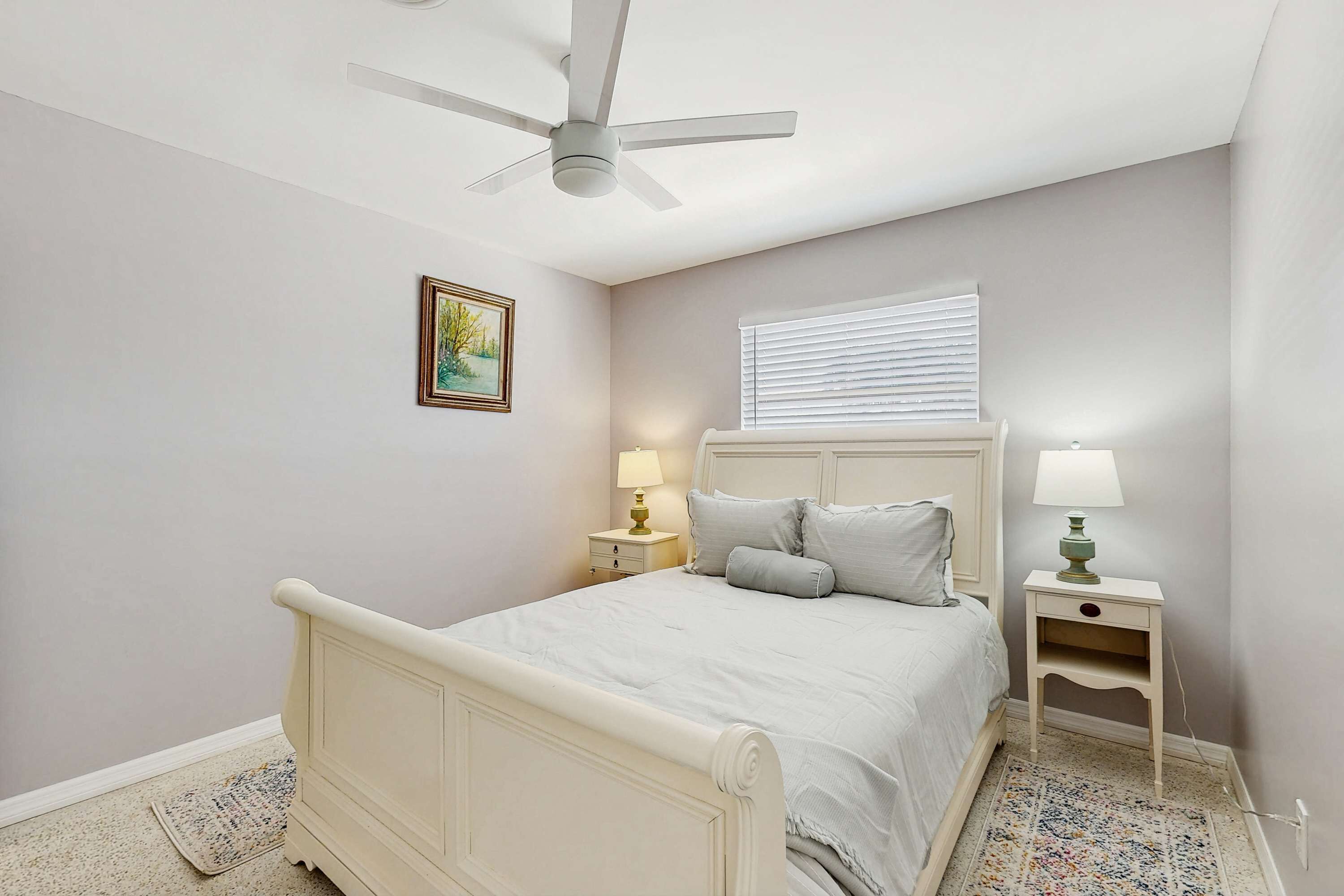 Une chambre à coucher comprenant un lit, une table de chevet, une lampe, un ventilateur de plafond, une fenêtre avec des stores et un tableau encadré accroché au mur.