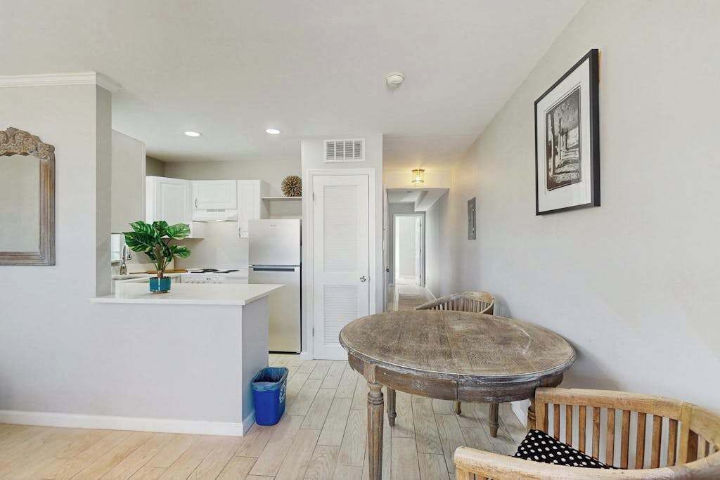 A modern kitchen with white cabinets, a dining area with a round table and wooden chairs, and a hallway leading to another room.