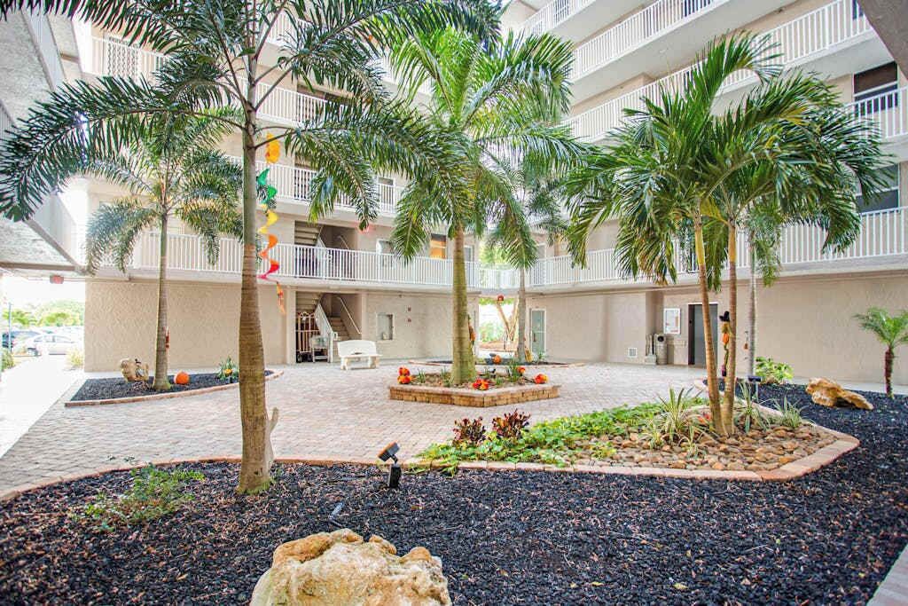 A courtyard with palm trees, a central planter with flowers, and a building with balconies.