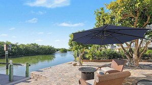 A patio with wicker furniture, a table, and a large umbrella overlooking a river. - Immaculate bay front home w/in ground pool, spa, paver patio & porch (Bonita Springs)
