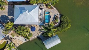 An aerial view of a property with a large metal roofed structure, a swimming pool, and surrounding greenery. - Immaculate bay front home w/in ground pool, spa, paver patio & porch (Bonita Springs)