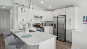 A modern kitchen with white cabinets, stainless steel appliances, and a central island with blue placemats. - Immaculate bay front home w/in ground pool, spa, paver patio & porch (Bonita Springs)