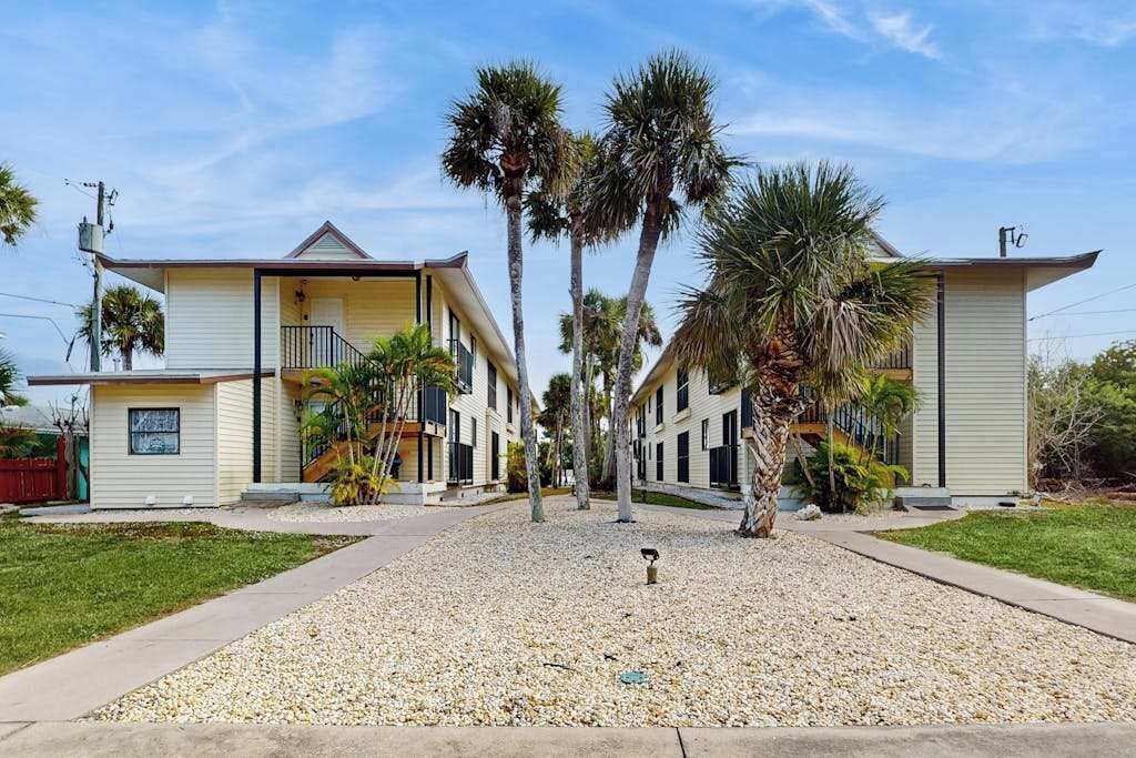 A row of two-story buildings with a gravel driveway and palm trees.