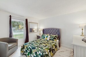 A bedroom with a bed, a chair, a nightstand, a lamp, and a window with curtains.