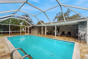 Una piscina con cerramiento de vidrio, paredes de piedra y mobiliario exterior.