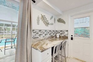 Una cocina con encimera de mármol, armarios blancos, un comedor con sillas y una piscina visible a través de un ventanal.