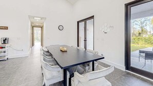 A modern dining area with a dark table and white chairs, a clock on the wall, and a view through a glass door. - Custom-built home with private patio, central AC, & W/D - dog-friendly (Port Charlotte)
