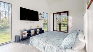 A bedroom with a bed, a TV mounted on the wall, a sliding glass door to a garden, and a small shelf with decorative items. - Custom-built home with private patio, central AC, & W/D - dog-friendly (Port Charlotte)