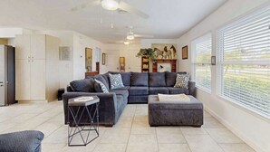 A spacious living area with a large dark-colored sofa, a smaller sofa, and a coffee table. There are ceiling fans, a bookshelf with decorative items, and a window with blinds. - Spacious coastal Pet friendly home close to beaches and shopping. (Port Charlotte)