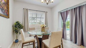 Zona pranzo con tavolo in vetro, sedie, lampadario, finestra con persiane e una pianta.