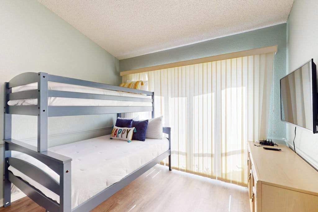 Un lit superposé avec un matelas, des oreillers et une pancarte où est écrit « JOYEUX ». Il y a une télévision posée sur un meuble en bois et une fenêtre avec des stores.
