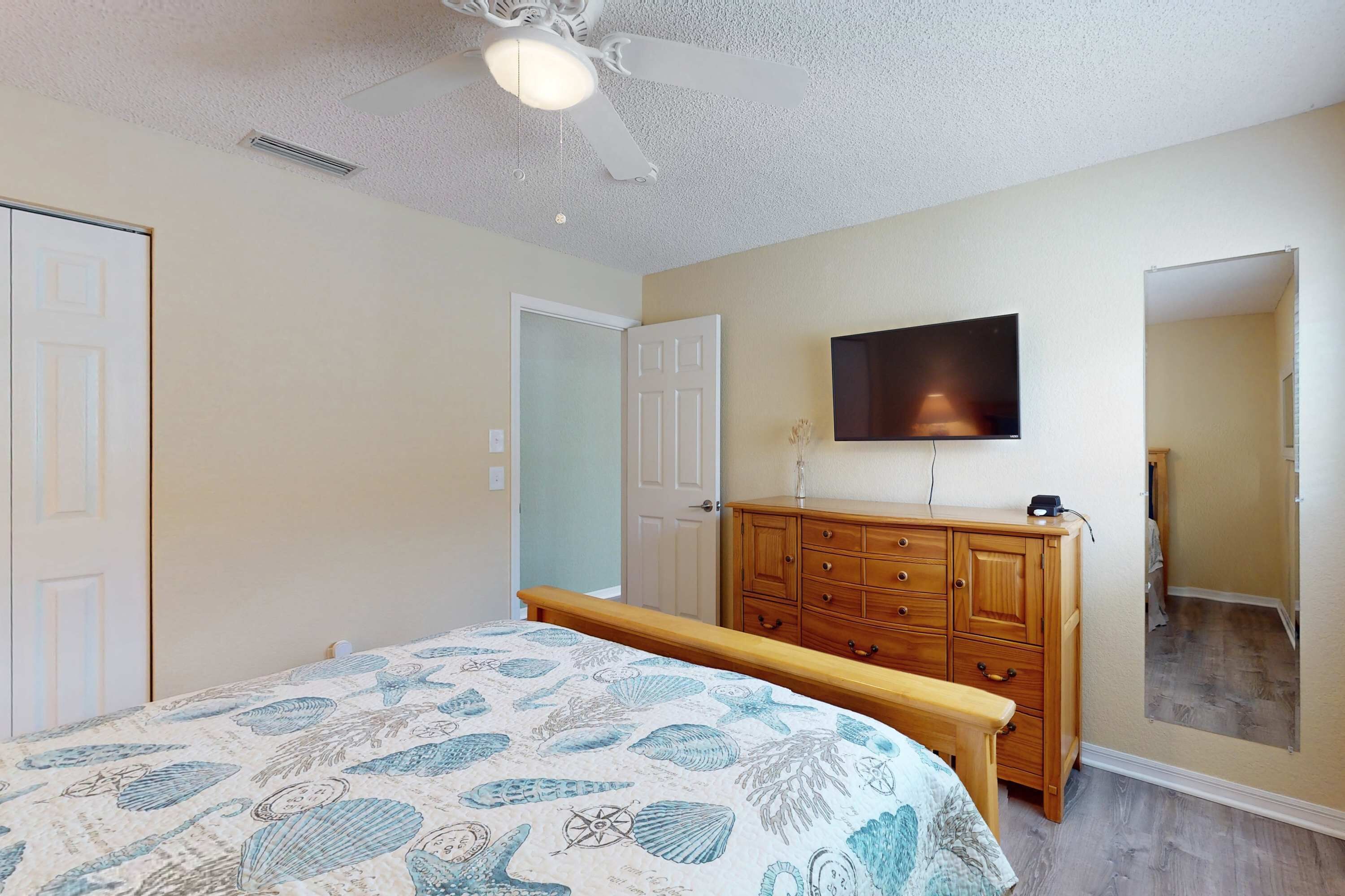 A bedroom with a bed, a wooden dresser, a ceiling fan, a TV mounted on the wall, and a door.