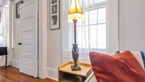 A room with a wooden floor, a white door, a lamp on a wooden table, and a red cushion on a white sofa. - Casa Blanca|Near Ft Benning|Pet Friendly|King Bed (Columbus)