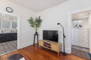 Een woonkamer met een televisie, een plant, een lamp, en een keuken zichtbaar door een open deur.