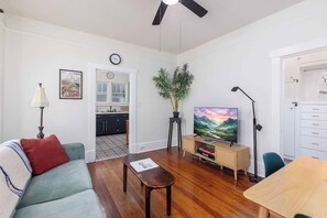 Una sala de estar con sofá, mesa de centro, televisor, planta y cocina al fondo.