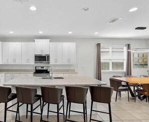 Une cuisine moderne avec des meubles blancs, un îlot central doté de tabourets, une table à manger avec des chaises et une grande fenêtre avec des stores.