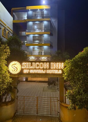 Un edificio de varios pisos con balcones, un letrero de neón que dice 'Silicon Inn by Suviraj Hotels' y una entrada con portón.