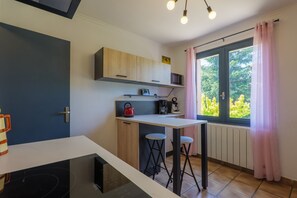 Een compacte keuken met een magnetron, waterkoker en koffiezetapparaat op een wit aanrecht, een klein raam met gordijnen, en een deur met een handvat.