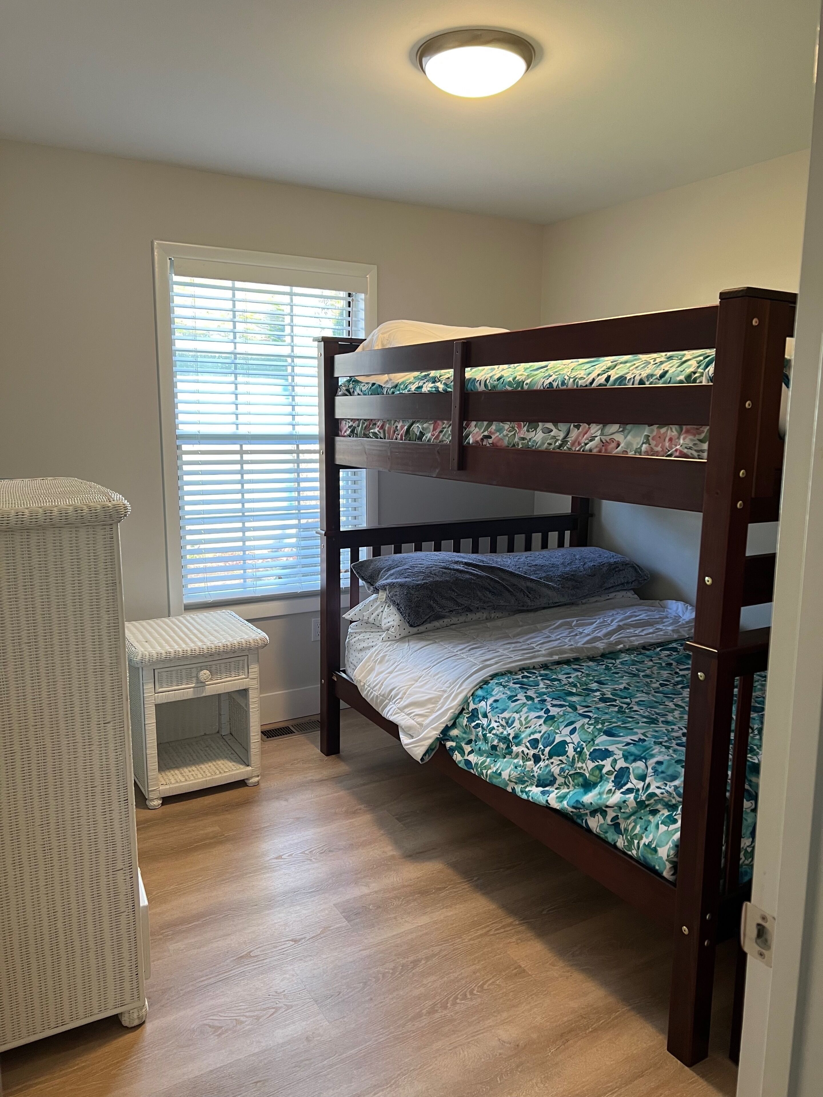 A bunk bed with a floral bedspread, a small white wicker side table, and a window with blinds.
