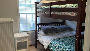 A bunk bed with a floral bedspread, a small white wicker side table, and a window with blinds.
