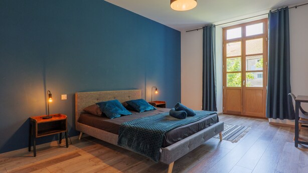 Une chambre à coucher avec un lit, des tables de chevet, une lampe, une fenêtre avec des rideaux et une vue sur l’extérieur.