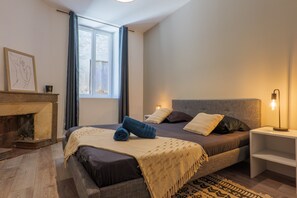 Un dormitorio con cama, mesita de noche, lámpara, chimenea y ventana.
