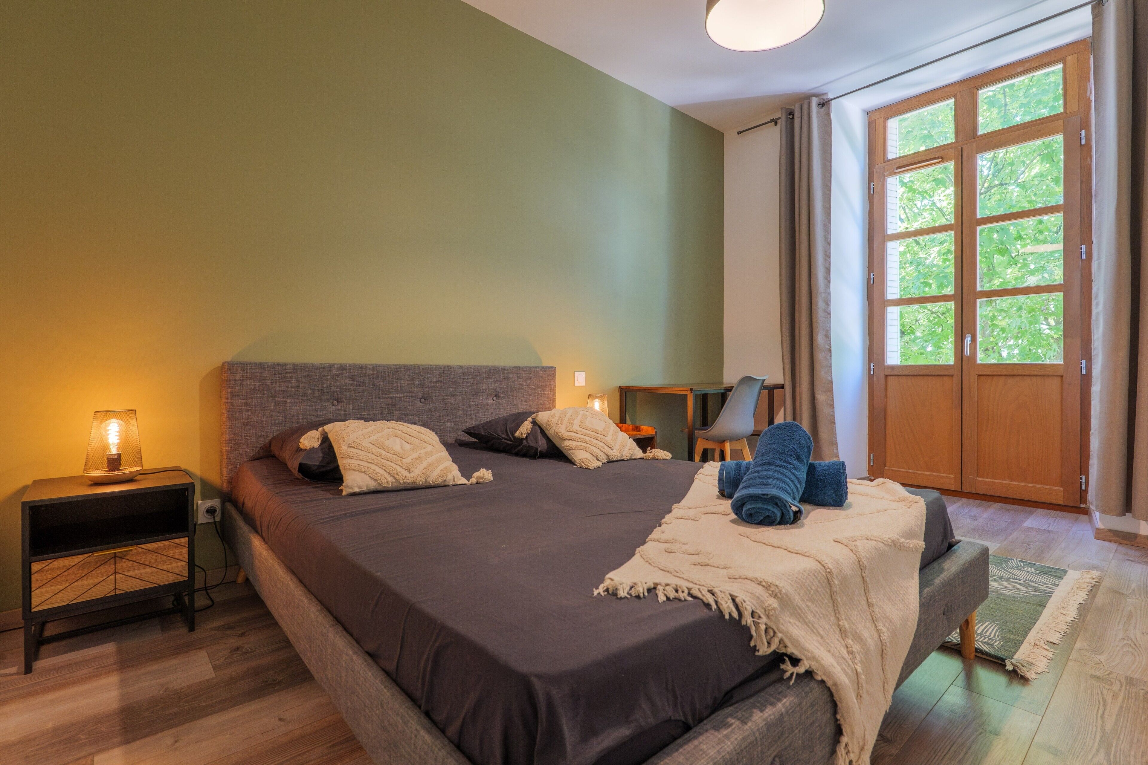 Ein Schlafzimmer mit Bett, Nachttischen, einer Lampe, einem Sessel und einem großen Fenster.