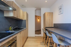 A modern kitchen with wooden cabinets, a black countertop, and a built-in oven. There are two bar stools and a wall-mounted light fixture.