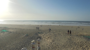 Uma extensa praia de areia com pessoas caminhando e em pé, céu claro e o oceano ao fundo.