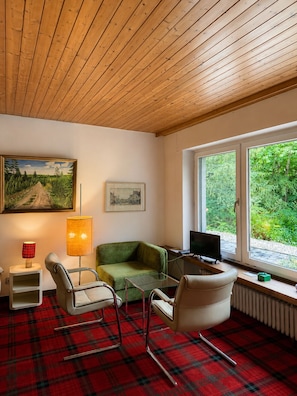 A room with a wooden ceiling, a green sofa, a white chair, a red plaid carpet, a lamp, a painting, and a window with a view of greenery.