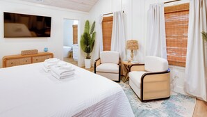 A hotel room with a large bed, a television, a chair, a small table with a lamp, and a window with curtains. - Palmwood Haven | Private Pool, 2nd House From Sand! (Tybee Island)