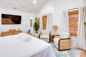 A hotel room with a large bed, a television, a chair, a small table with a lamp, and a window with curtains. - Palmwood Haven | Private Pool, 2nd House From Sand! (Tybee Island)