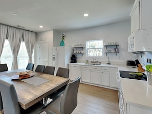 A modern kitchen with white cabinets, a dining table with chairs, and a window with a view of palm trees. - Limetime Tybee | Vibrant, Upgraded, Outdoor Living (Tybee Island)