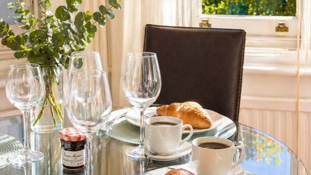 Une table dressée avec des verres à vin, des assiettes et un croissant, donnant sur un paysage urbain et des arbres, vu à travers une fenêtre.