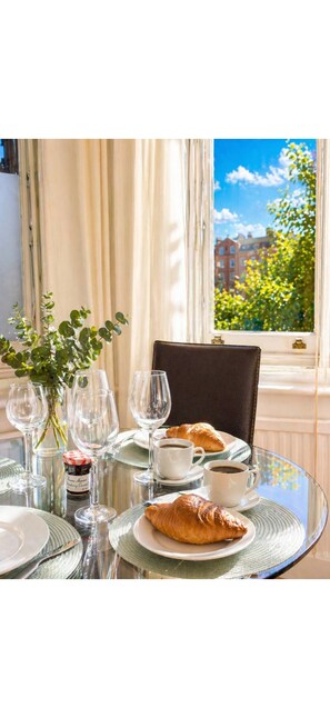 Ein Tisch ist gedeckt mit Weingläsern, Tellern und einem Croissant, mit Blick auf Gebäude und Bäume durch ein Fenster.