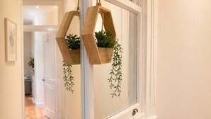 Un couloir avec une porte blanche, un miroir reflétant une pièce, et deux jardinières hexagonales en bois ornées de plantes vertes.