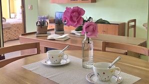 Una mesa de comedor con dos tazas y un florero, sillas de madera, y una zona de cocina con armarios al fondo.